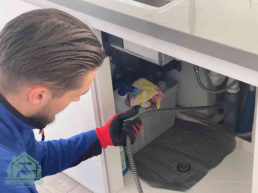 Een monteur voert een flexibele ontstoppingsveer in onder een keukenkastje om een verstopping voorbij de sifon te bereiken in Ede