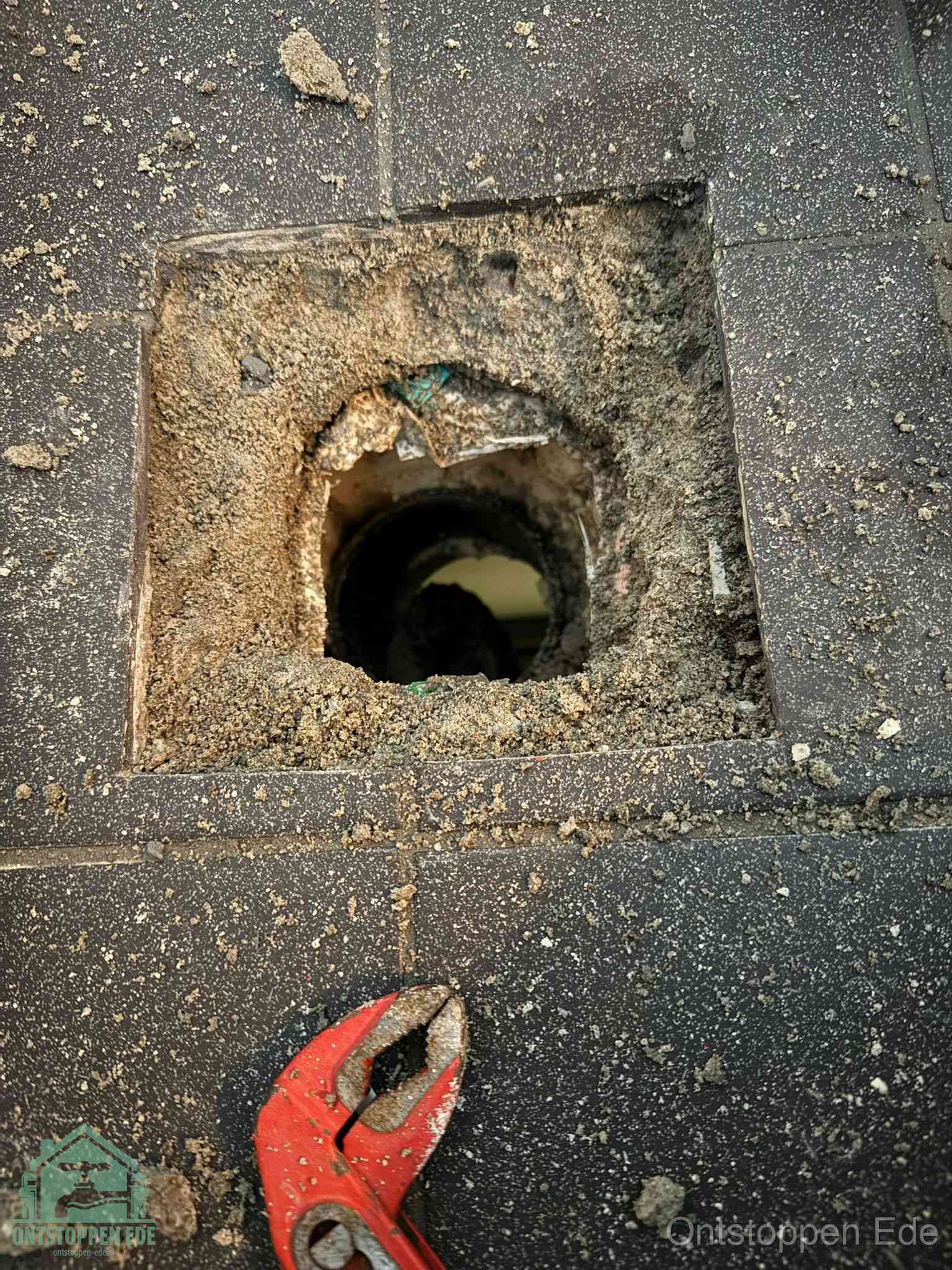 Een geopend rioolputje in een betontegelvloer met losgemaakt rooster en een zichtbare ronde afvoerbuis, het achterhalen waar de afvoerverstopping zit in Ede