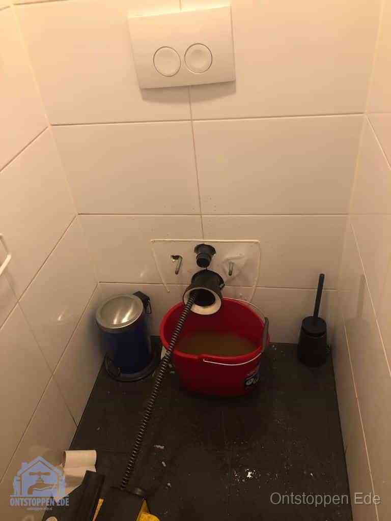 Een ontstopte wand-wc met verwijderde toiletpot waar een ontstoppingsveer in de wanduitgang verdwijnt, het achterhalen waar de verstopping zit in Ede
