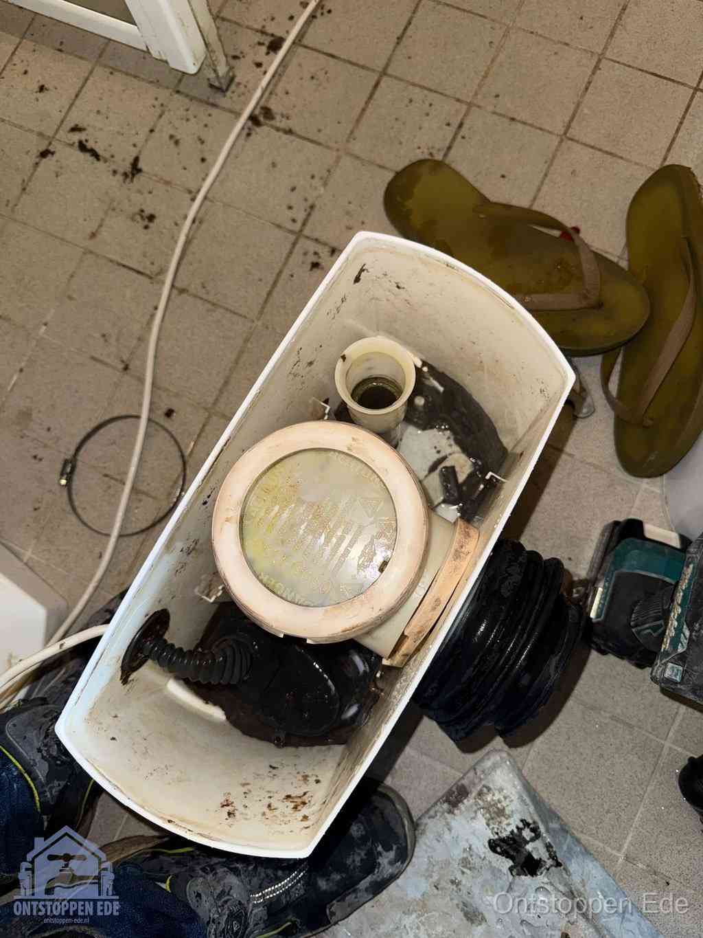 Een gedemonteerde toiletpot op de vloer met ontstoppingsveer en gereedschap ernaast, het verwijderen van een vastzittend voorwerp uit de wc in Ede